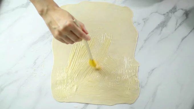 打ち粉を叩いた台の上にタルト生地を置き、めん棒で25x35cmほどの長方形にのばします。生地を縦長に置きます。
ハケなどを使って上面一帯に薄くバターを塗ります。