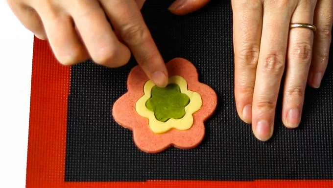 Place the dough on a baking sheet lined with parchment paper (or use a silicone baking mat). Start by laying down the largest flower-shaped dough, then use a smaller flower-shaped cookie cutter to cut out the center. Fill the empty space with a different colored dough that is the same size as the cut-out flower. Then, using an even smaller flower-shaped cookie cutter, cut out the center of the second layer, and place a different colored dough of the same size in the hollow space. Repeat this process with smaller flower shapes until you have completed the design.