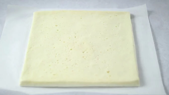 ロールケーキ生地の上下のラップまたはオーブンペーパーを剥がし、好みの色味の方が外側にくるように下側におき、新しいオーブンペーパーを敷きます。